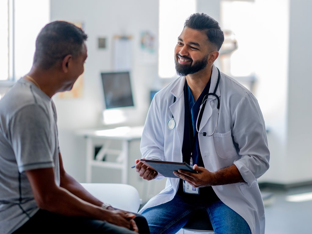 A patient sits up on an exam table as he talks with his male doctor about his health concerns. The doctor is dressed professionally and taking notes electronically.