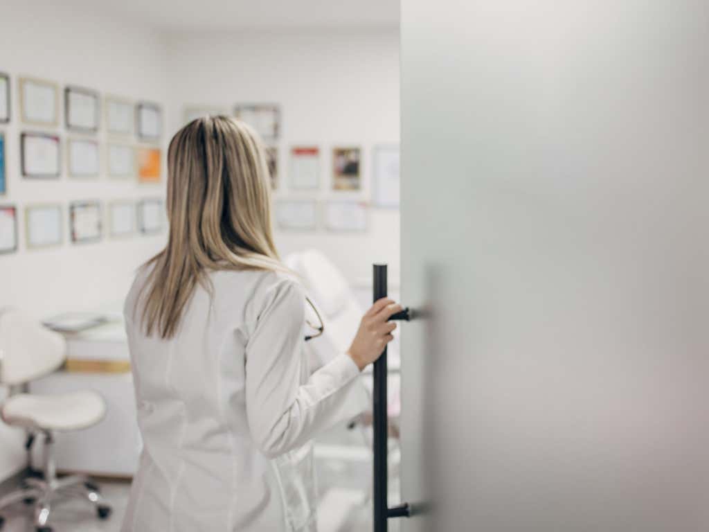 Female doctor cosmetologist in lab coat opening door at the beauty clinic.